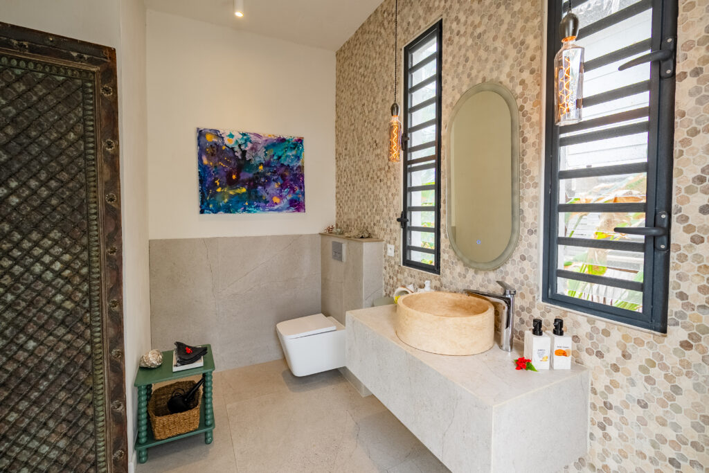 Oceanfront Hideaway Suite bathroom with stone washbasin, illuminated mirror, mosaic tiles and tropical artwork.